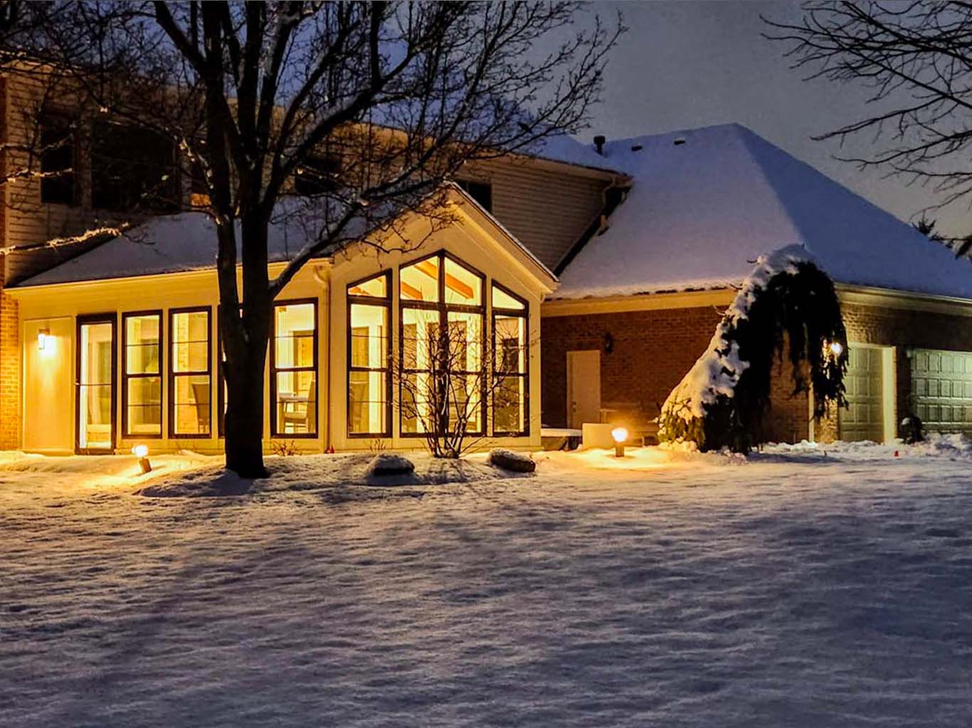 A house with a glowing sunroom, snow-covered ground, and illuminated landscape lighting at dusk.