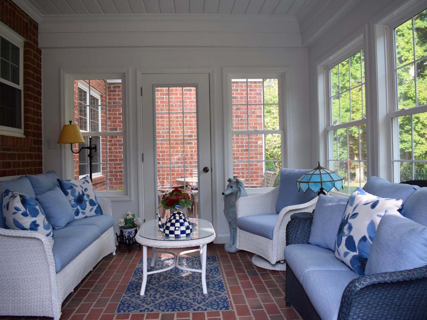 A sunroom with brick walls features two wicker sofas with blue cushions, a glass coffee table with a checkered vase, a floor lamp, and a large window offering a view of trees.