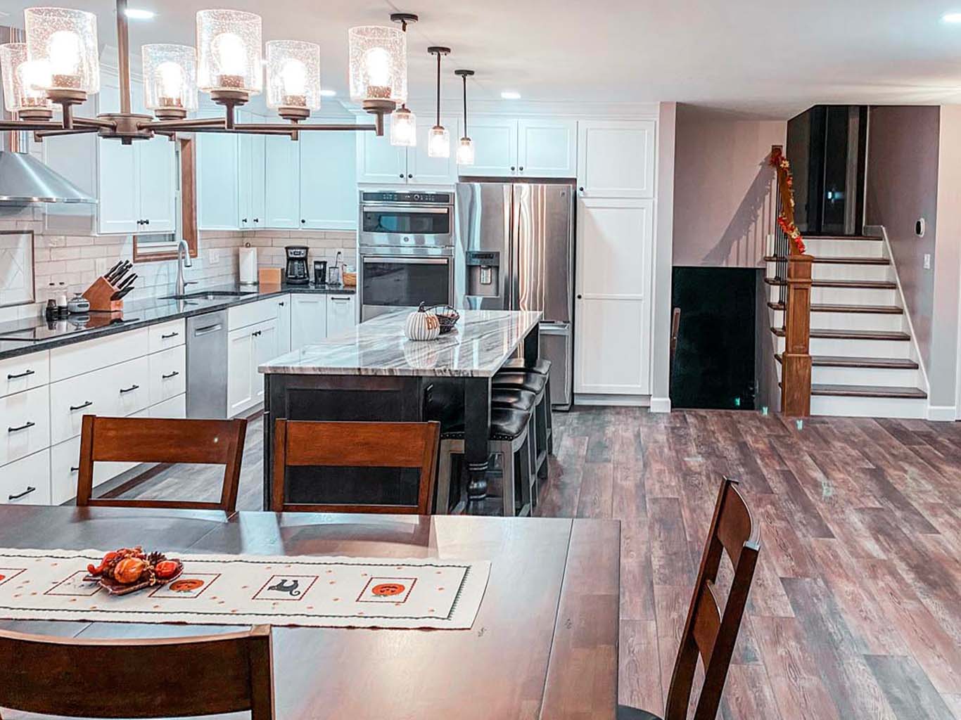 Modern kitchen and dining area with wooden floors, white cabinets, stainless steel appliances, a kitchen island with stools, and a staircase in the background.