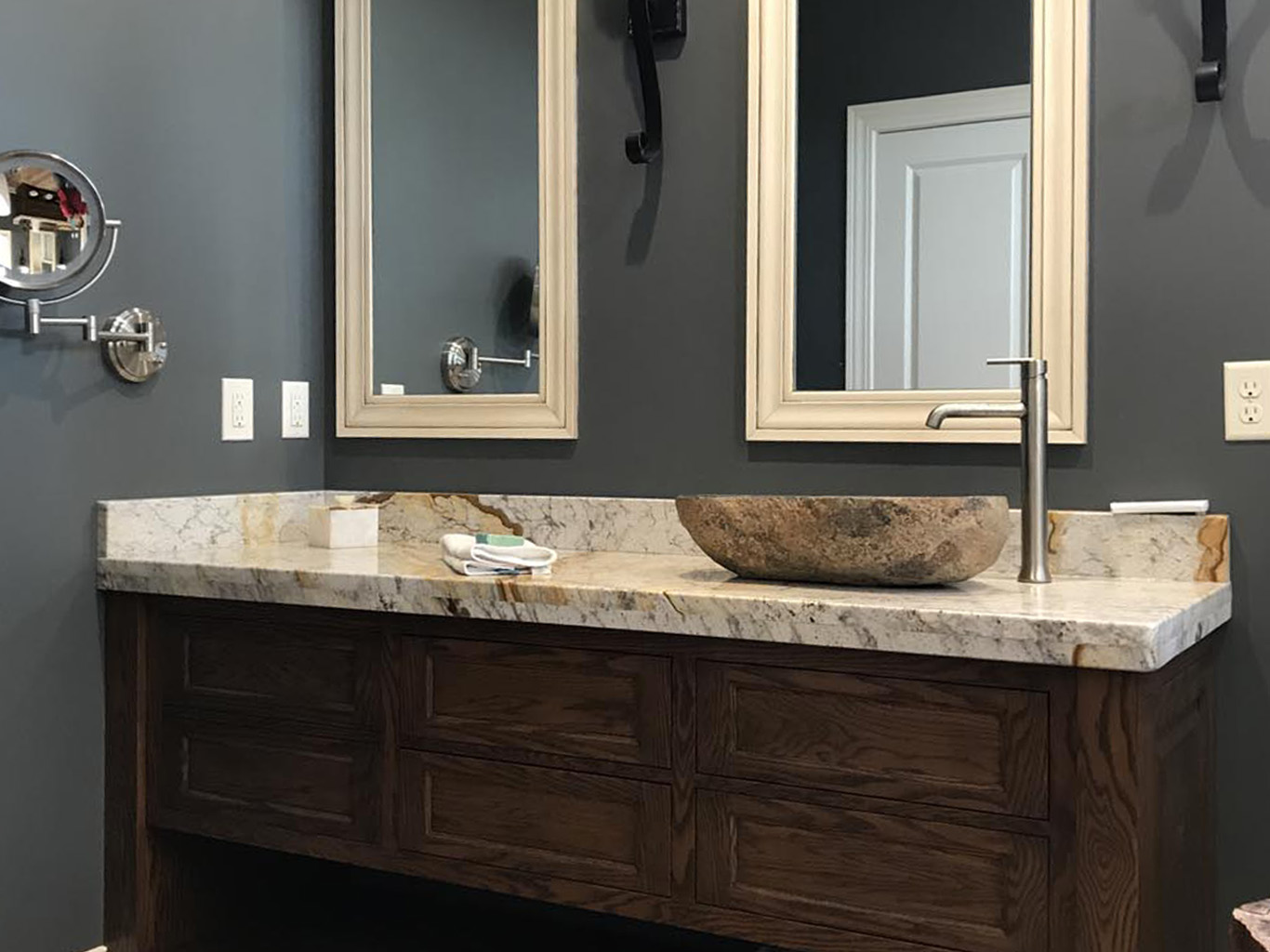 Bathroom with a dark wood vanity, a marble countertop, a stone basin, and two mirrors. Dark gray walls and a mounted round mirror are also visible.