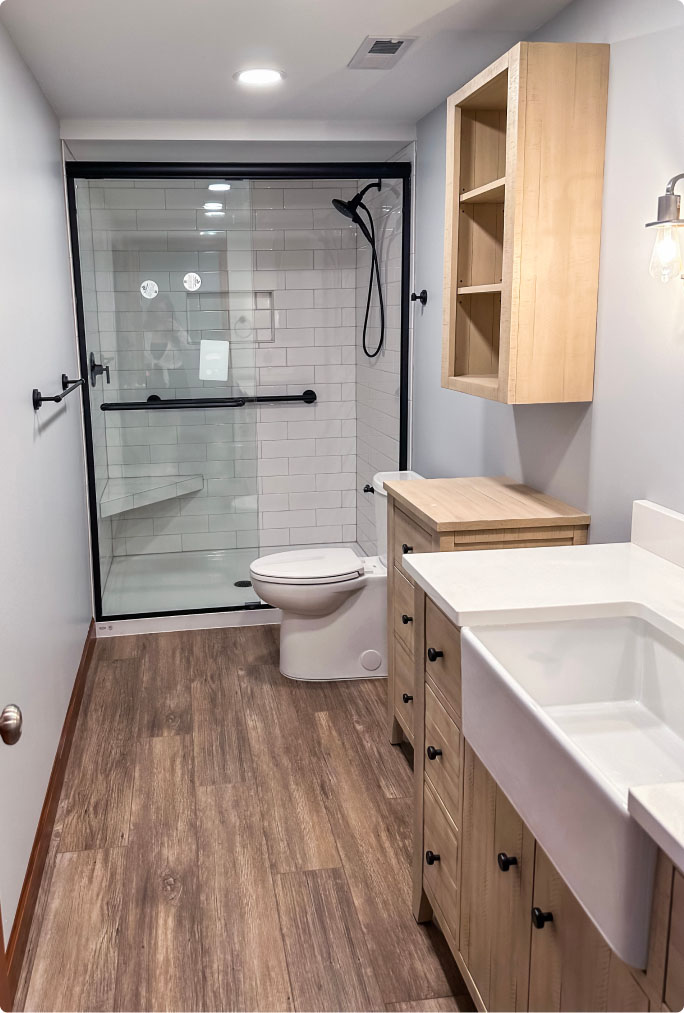 Modern bathroom with a shower, white toilet, and a sink cabinet. Wood flooring and cabinetry add warmth. A glass partition separates the shower area. Lighting fixture on the wall.