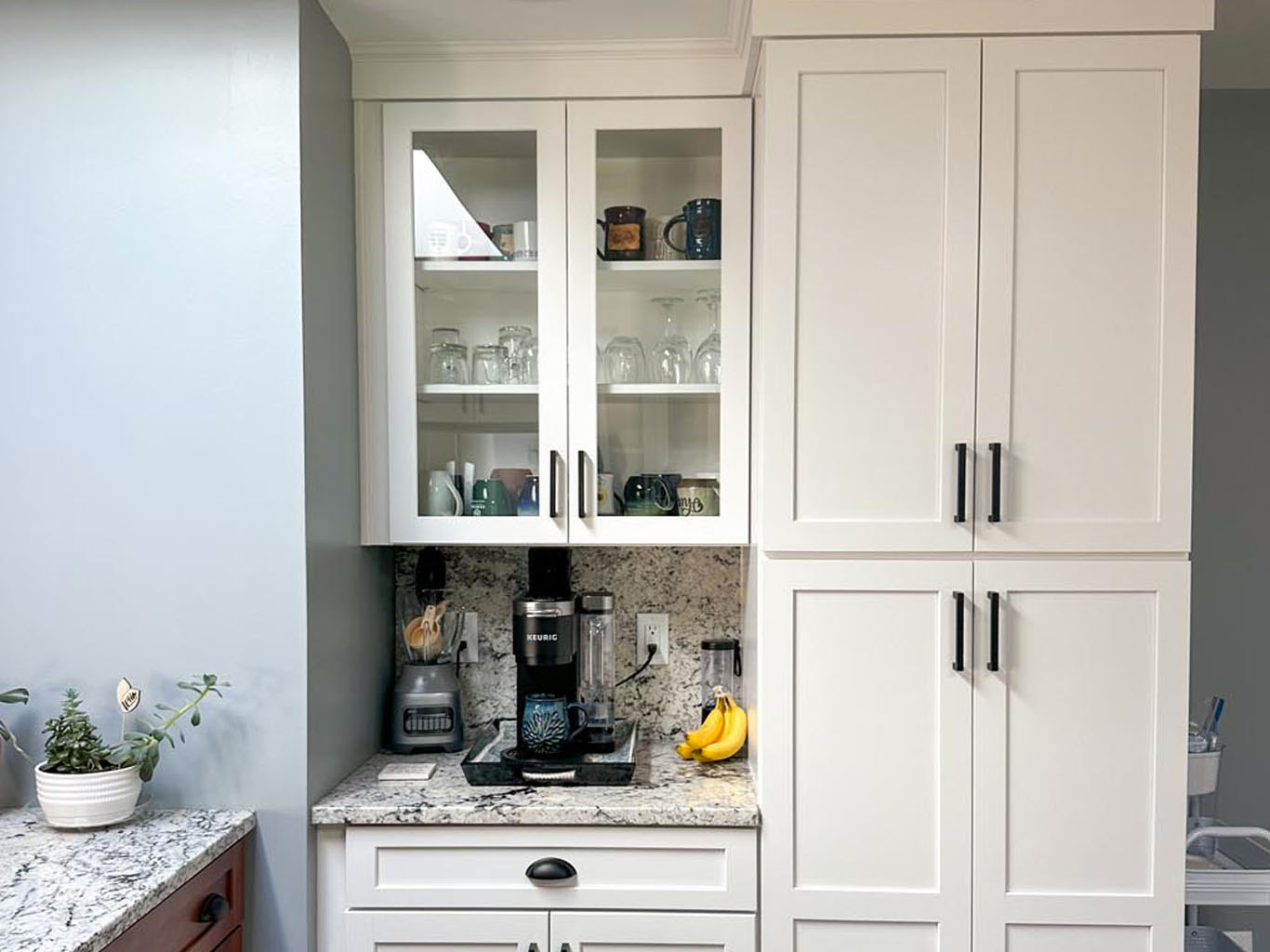 White kitchen cabinets with glass doors display mugs and glasses. A coffee maker and blender sit on the countertop, along with a bunch of bananas and a potted plant.