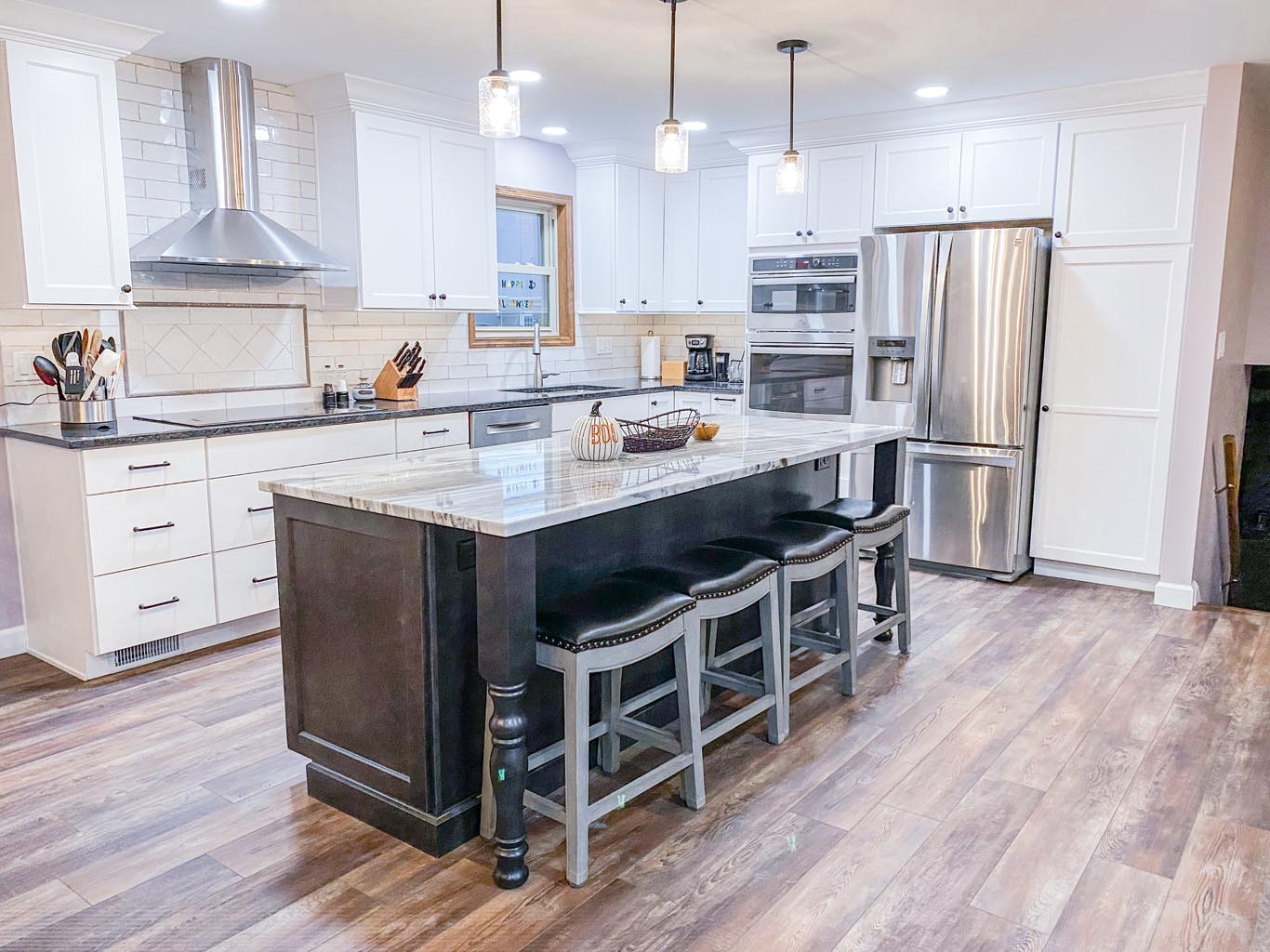 Modern kitchen with white cabinets, stainless steel appliances, and a large island with dark base. Three pendant lights hang above, and four stools are neatly arranged at the island.