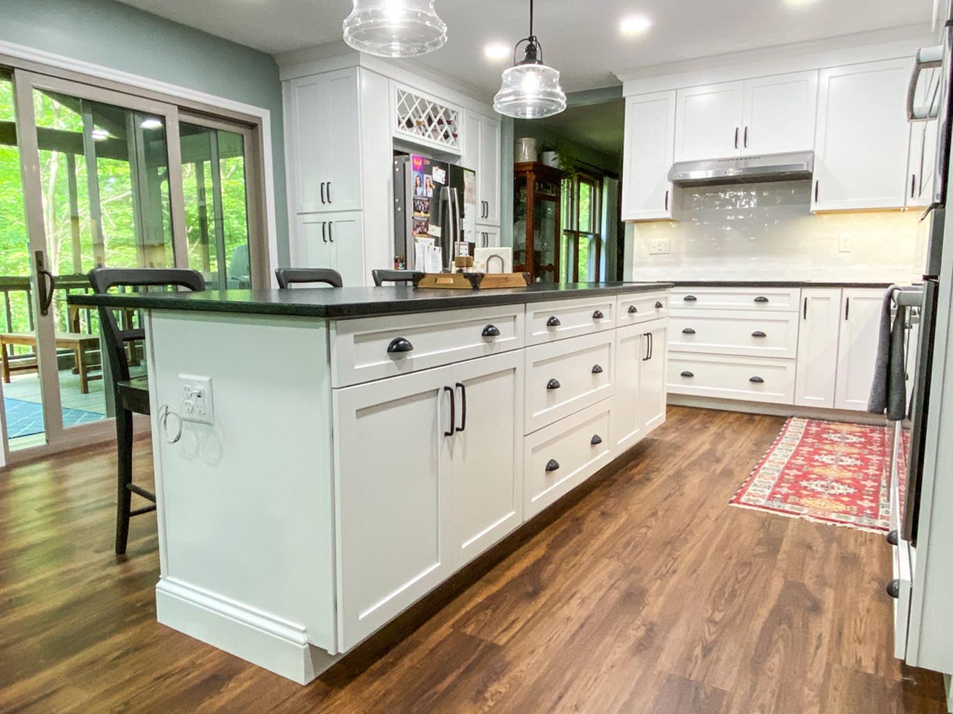 Spacious kitchen with white cabinets, dark countertops, and hardwood floors. Pendant lights hang above the island, and a sliding glass door leads to a greenery-filled patio.