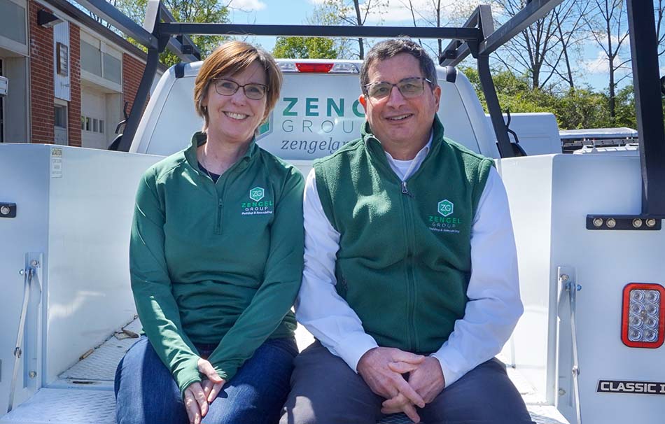 Two people sitting in the back of a pickup truck, wearing green Zengel Group jackets, smiling at the camera.