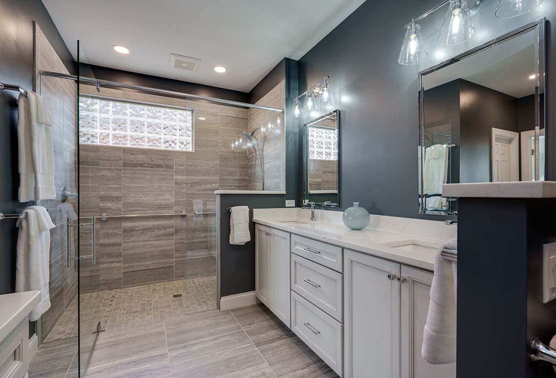 Modern bathroom with glass shower, gray tiled walls, white cabinets, and a large mirror.