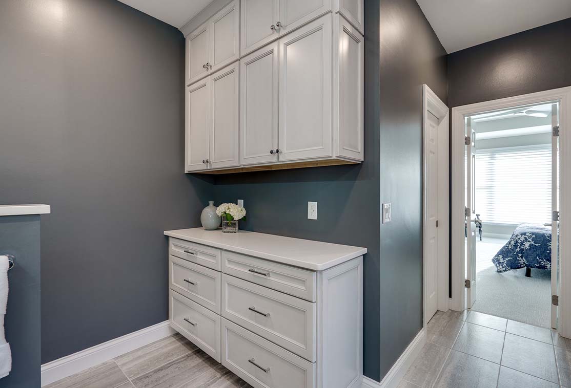 Modern bathroom with gray walls, white cabinetry, and silver hardware. A vase with flowers is on the counter. A doorway leads to a bedroom with natural light.