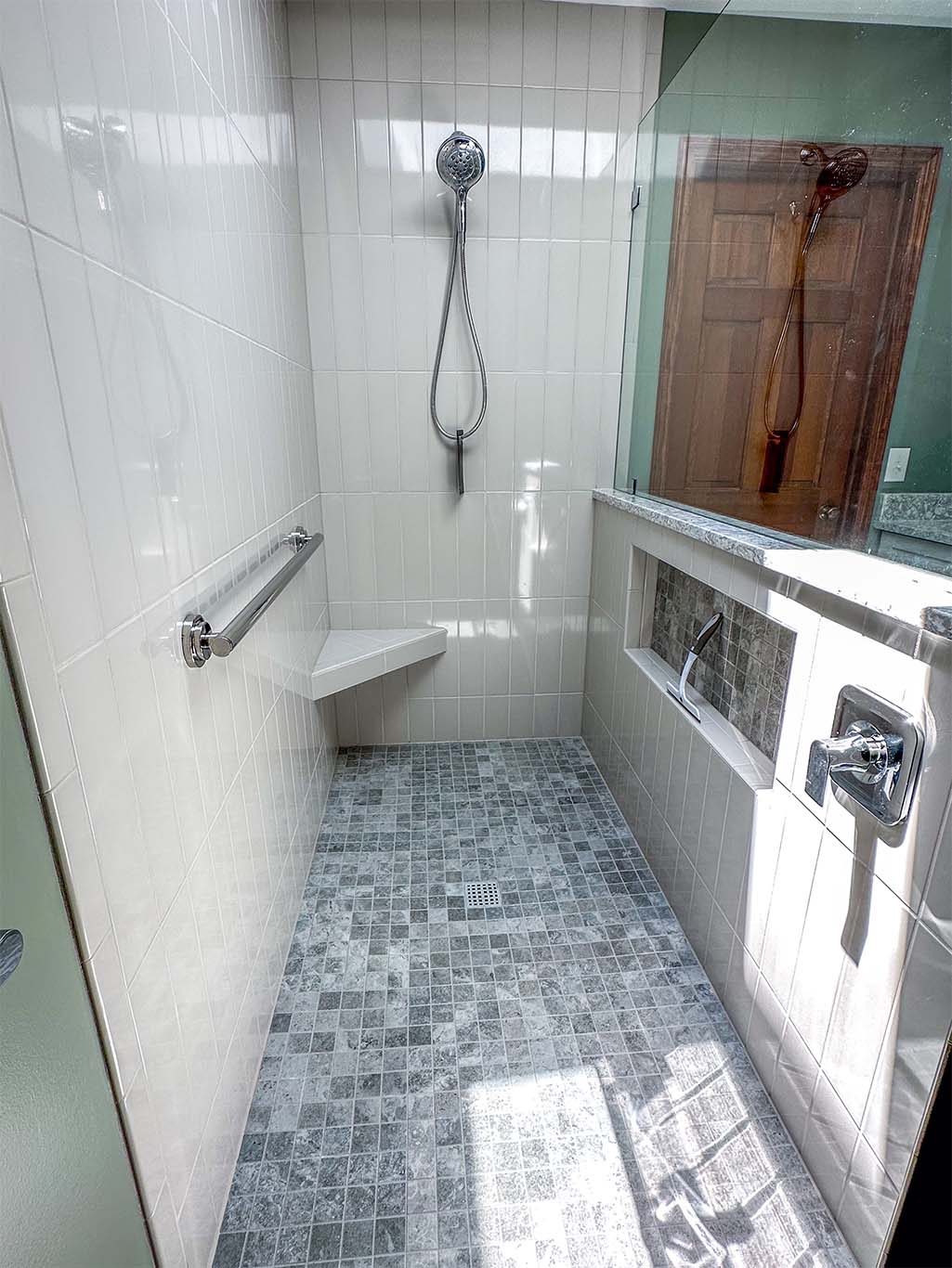 A small white-tiled shower with a corner seat, grab bar, handheld showerhead, and glass door. Light gray floor tiles and a built-in shelf are visible.