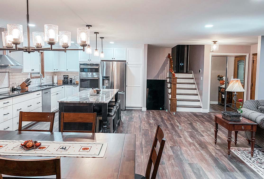 Open-concept kitchen and living area with hardwood floors, white cabinets, stainless steel appliances, a dining table, and a staircase leading upstairs.