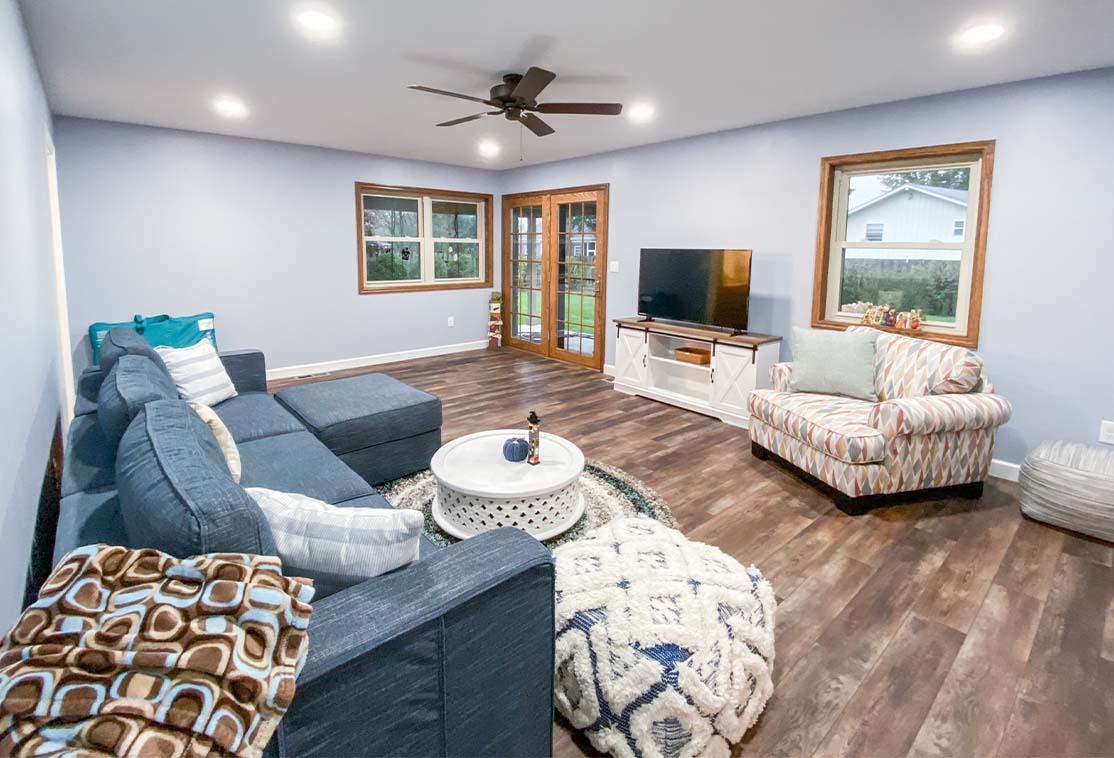 A living room with a gray sectional sofa, patterned armchair, round coffee table, TV on a stand, wooden floor, ceiling fan, and large windows.