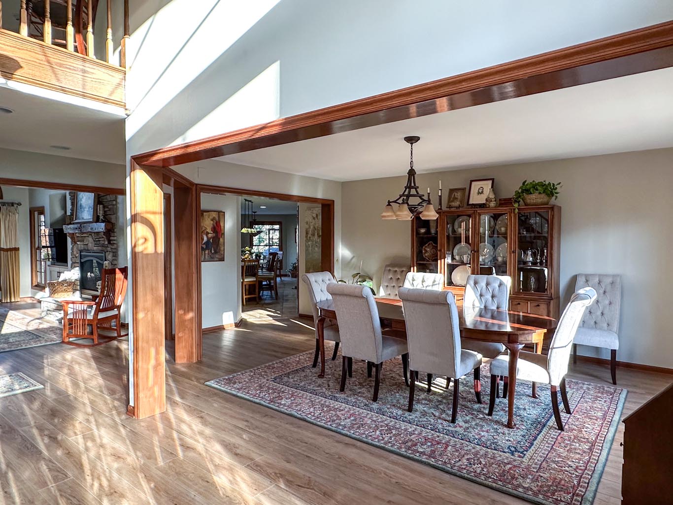 A whole house renovation in Dayton, Ohio that includes a bright dining room with a wooden table, eight upholstered chairs, a chandelier, and a china cabinet. Large windows allow ample natural light. A patterned rug is on the wooden floor.