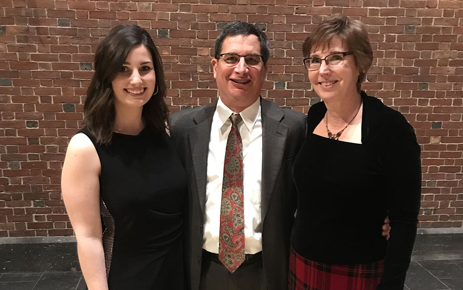 Three people, two women and one man, stand smiling in front of a brick wall. The man wears a suit, and the women are in formal attire.