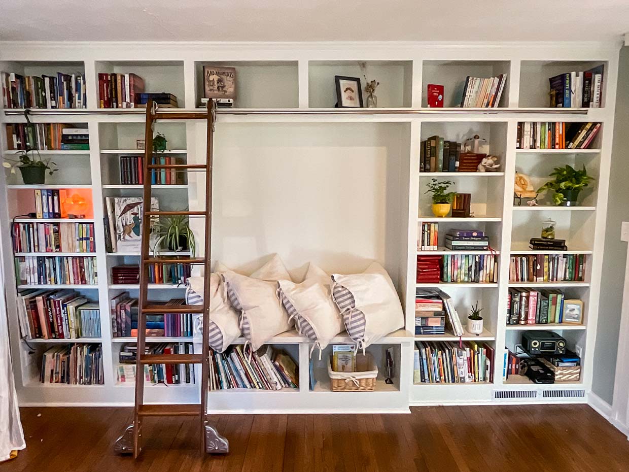 A large white bookshelf with various books, plants, and decorative items. A built-in bench with cushions is in the center, and a wooden ladder leans against the shelves.