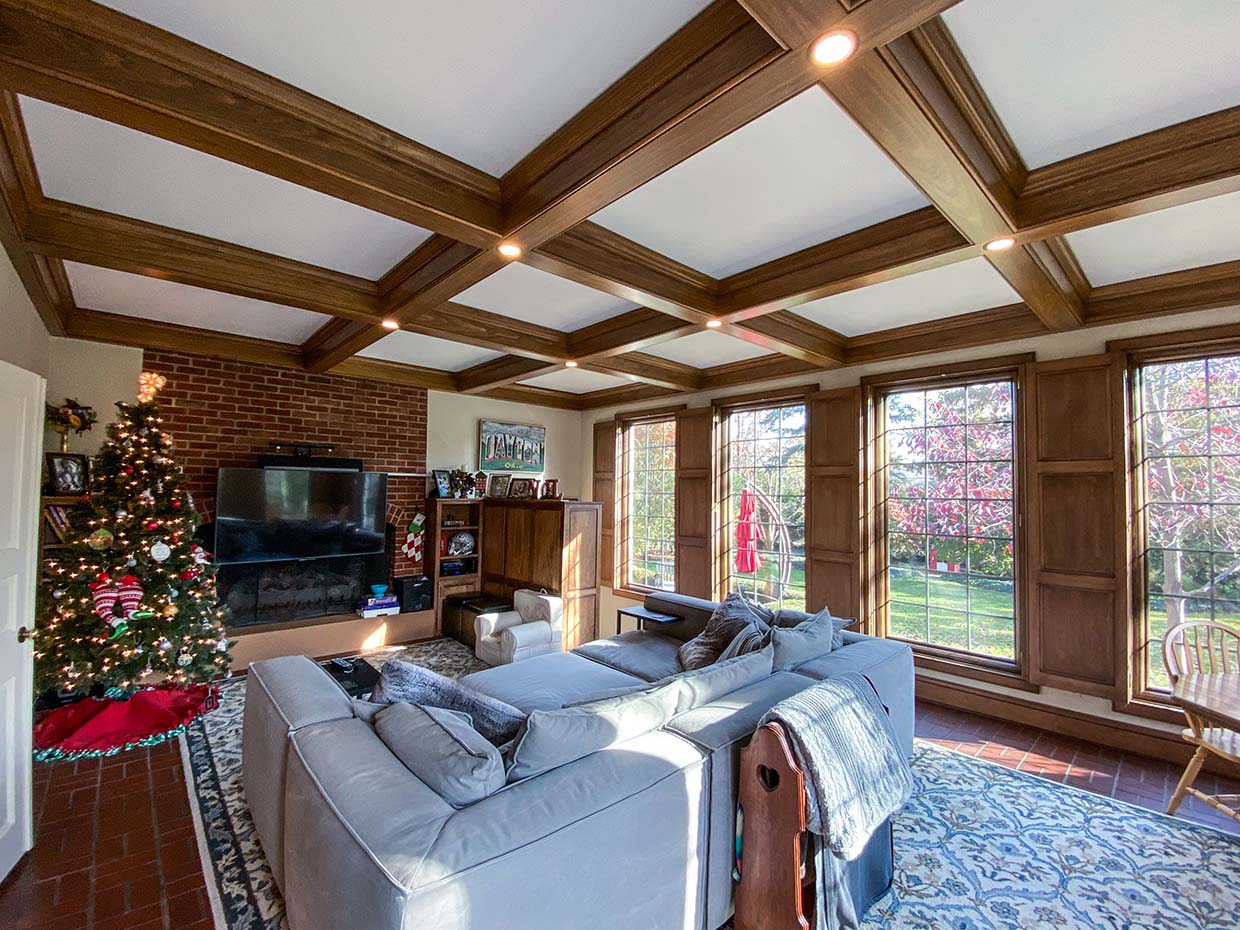 A living room with a decorated Christmas tree, large windows, a sectional sofa, and a TV on a brick wall. Natural light fills the space through the windows.