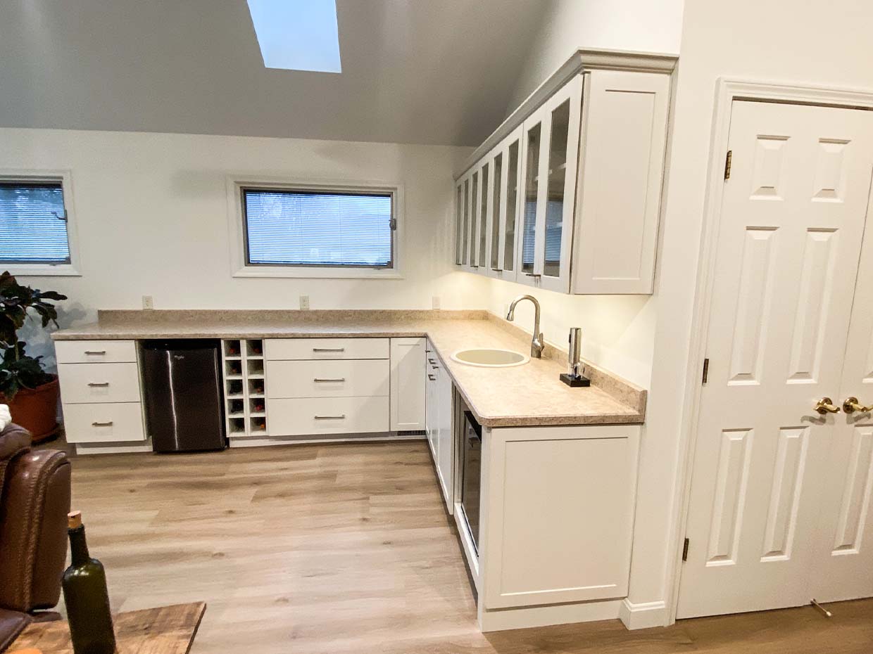 A modern kitchen with white cabinets, beige countertops, a sink, and a wine rack. Two small windows and a skylight provide natural light.