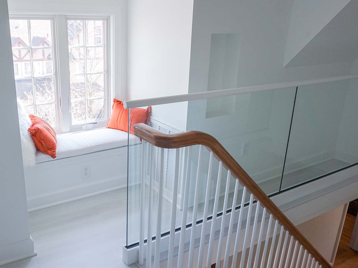 A bright corner with a window seat adorned with orange cushions. Glass and wood railing overlooks stairs. White walls and natural light create an airy atmosphere.