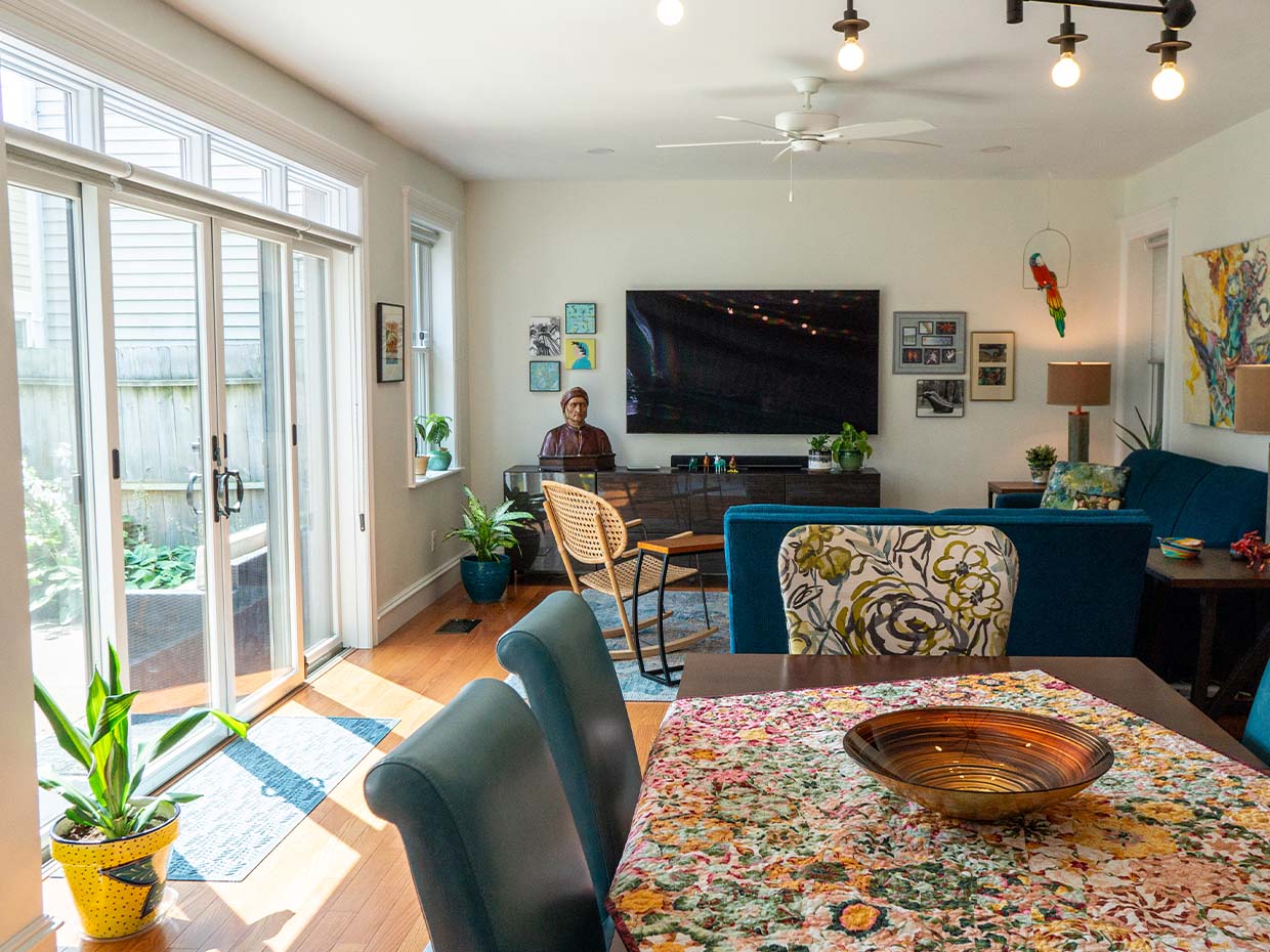 Bright living room with a dining area, large TV, and various artworks. A person sits at a desk by the window. Vibrant tablecloth and plants accent the space.