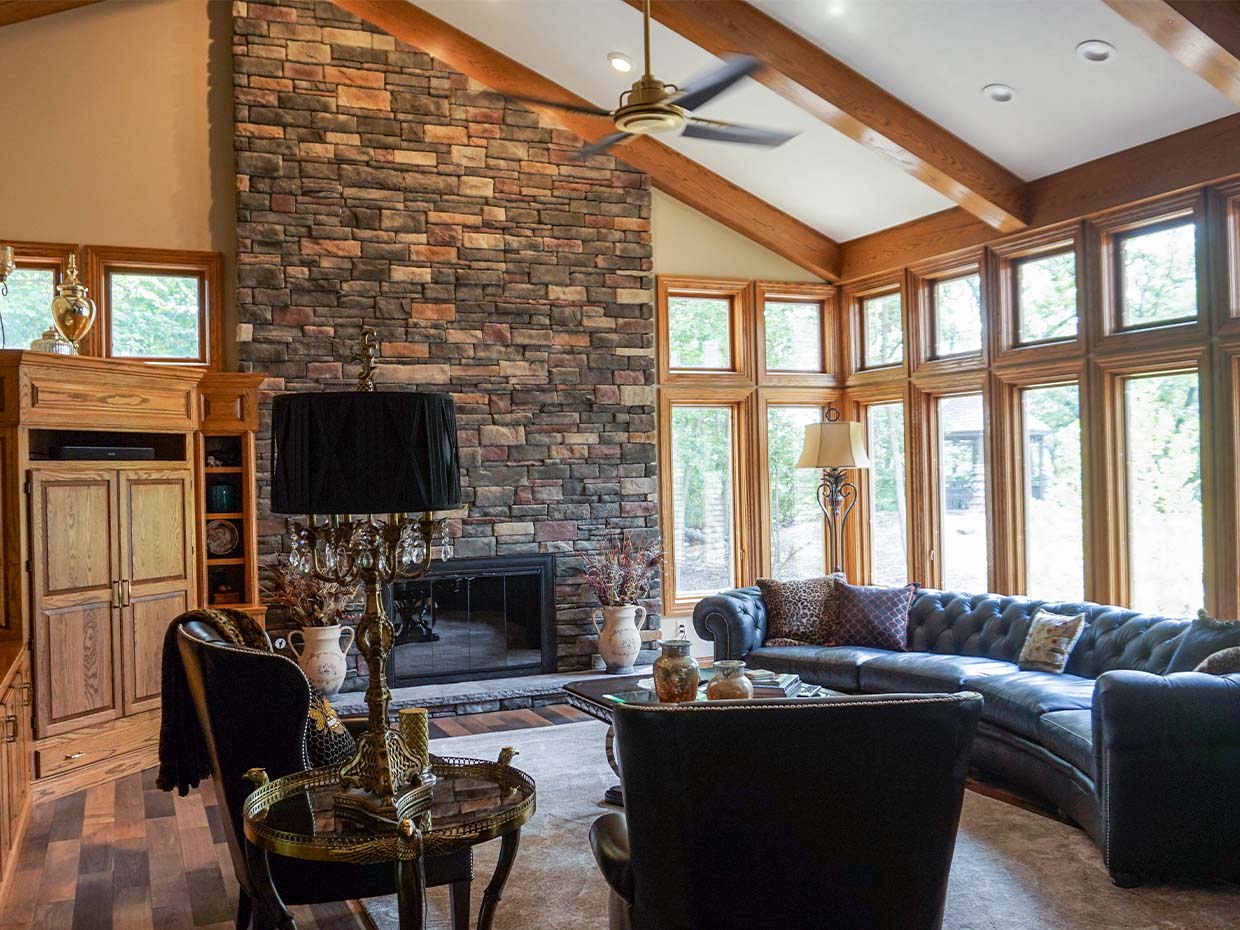 Living room with a high ceiling, stone fireplace, wooden beams, and large windows. Features a black leather sofa, two armchairs, and a decorative lamp on a side table.