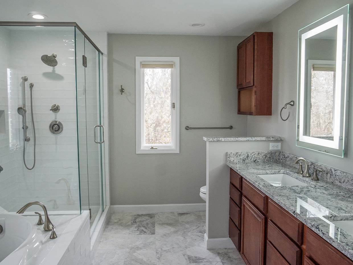 Modern bathroom with a glass shower, bathtub, granite countertop, and wooden cabinets. A window is on the wall above the toilet.