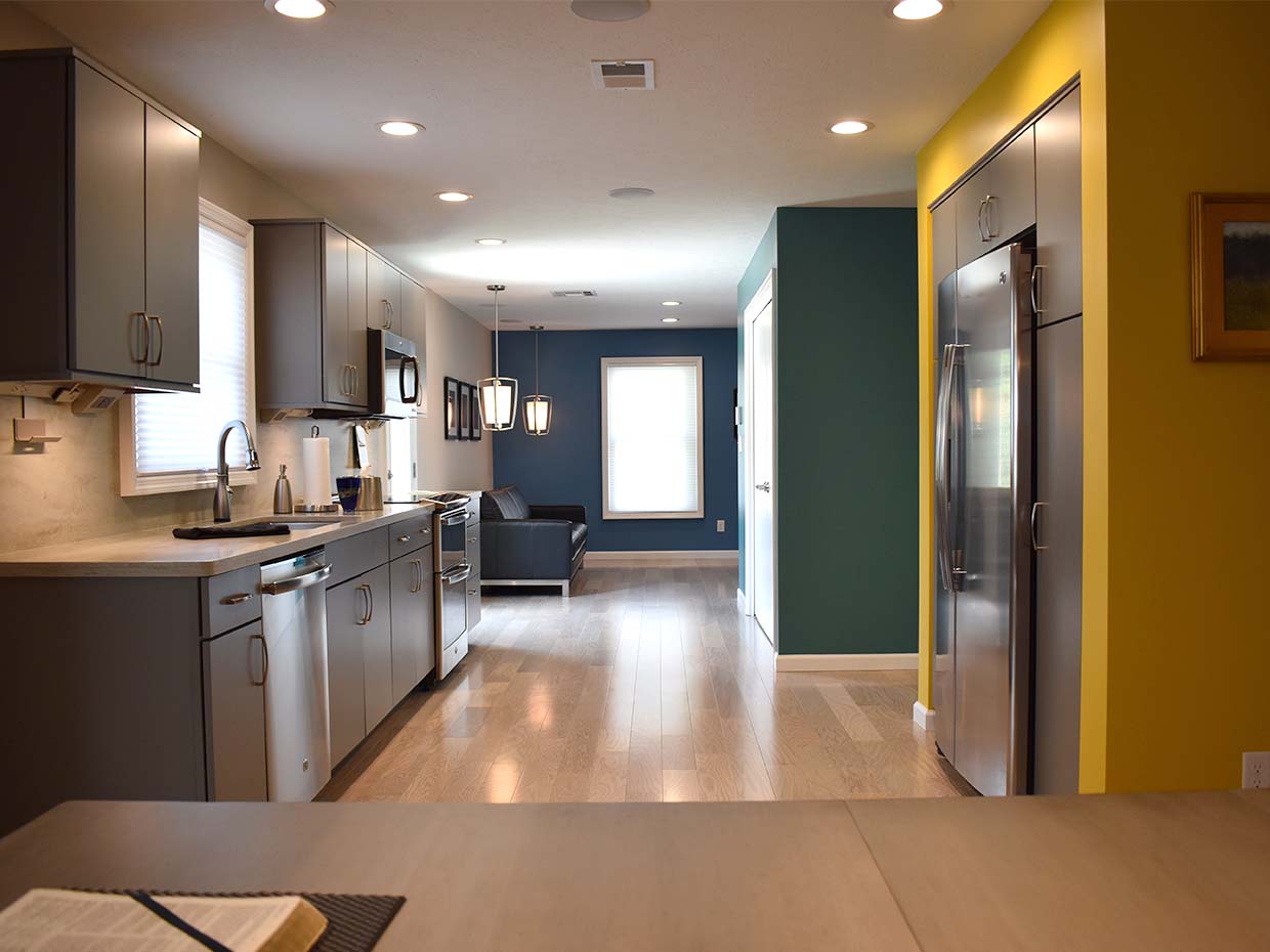 Modern kitchen view with gray cabinets, stainless steel appliances, and wooden flooring, leading to a living area with a dark sofa and teal accent wall.