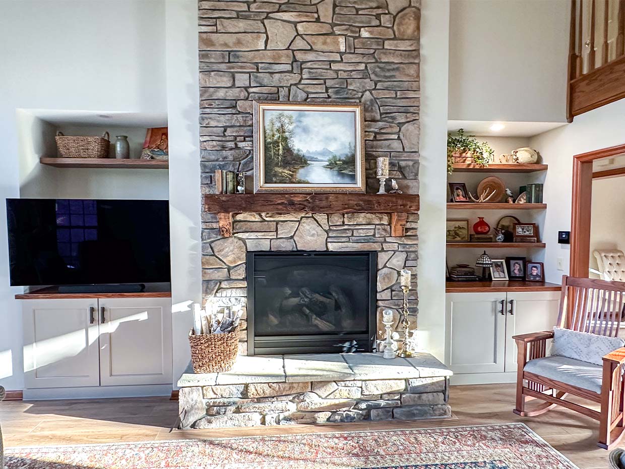 A cozy living room with a stone fireplace, framed artwork, and built-in shelves holding decor items. A TV is on the left, and a rocking chair is on the right. Natural light fills the space.