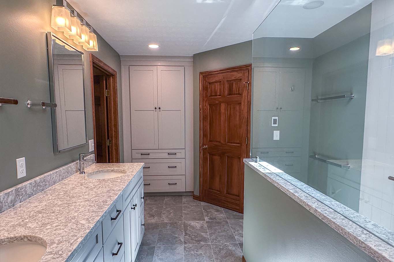 A bathroom with a granite countertop double vanity, large mirror, wooden cabinets, glass shower enclosure, and gray flooring.