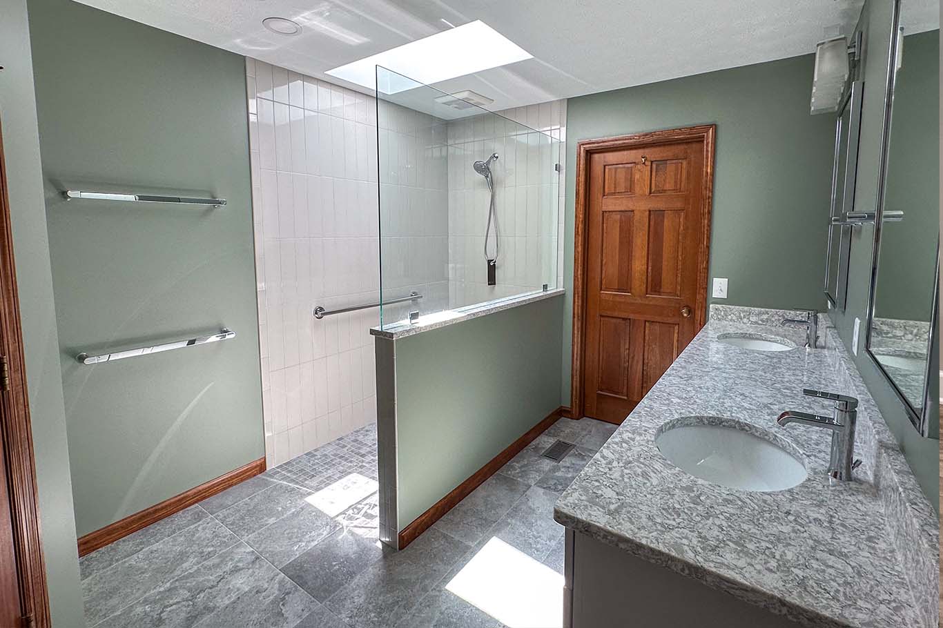 Modern bathroom with green walls, granite double sink counter, and a glass-enclosed shower. Wooden door and towel racks on the left wall. Skylight above.