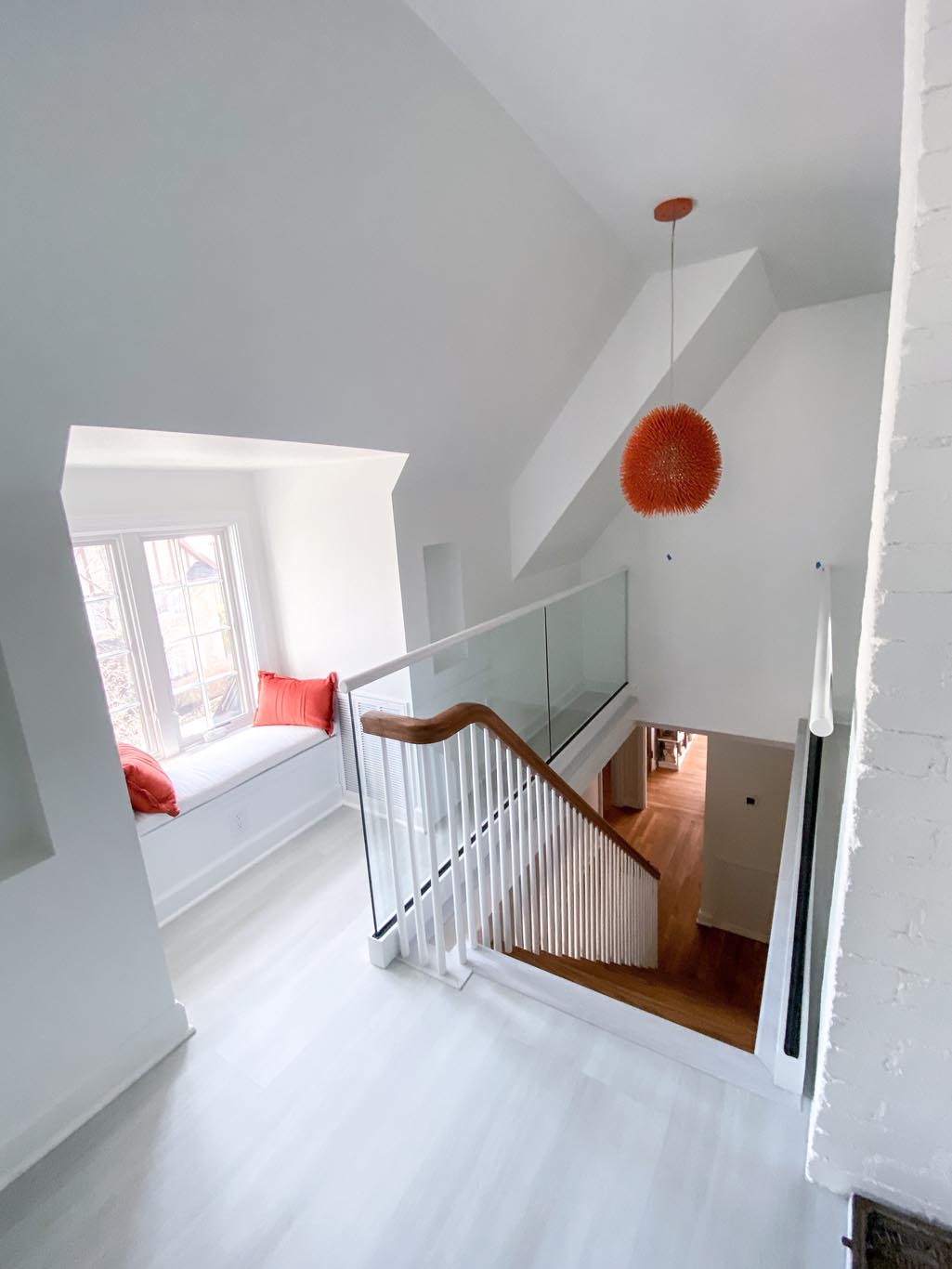 Bright white stairway with wooden handrail, glass panels, and an orange pendant light. A window seat with red cushions is on the left.