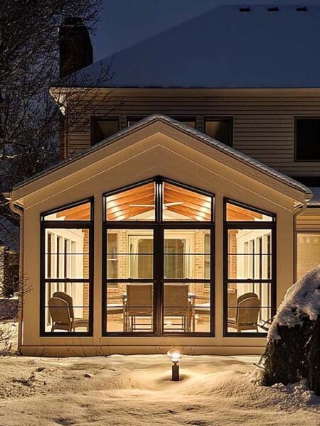 A warmly lit sunroom with large windows and a gabled roof is shown at night, surrounded by snow-covered ground.