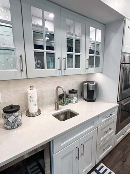 A modern kitchen corner with glass-front cabinets, a small sink, a coffee maker, and a countertop holding cups and a paper towel holder.