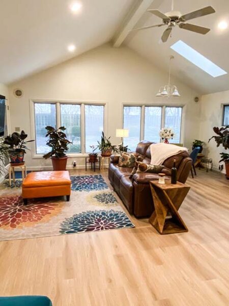 A bright living room with a leather couch, colorful rug, and an orange ottoman. Large windows, plants, and a ceiling fan are visible.