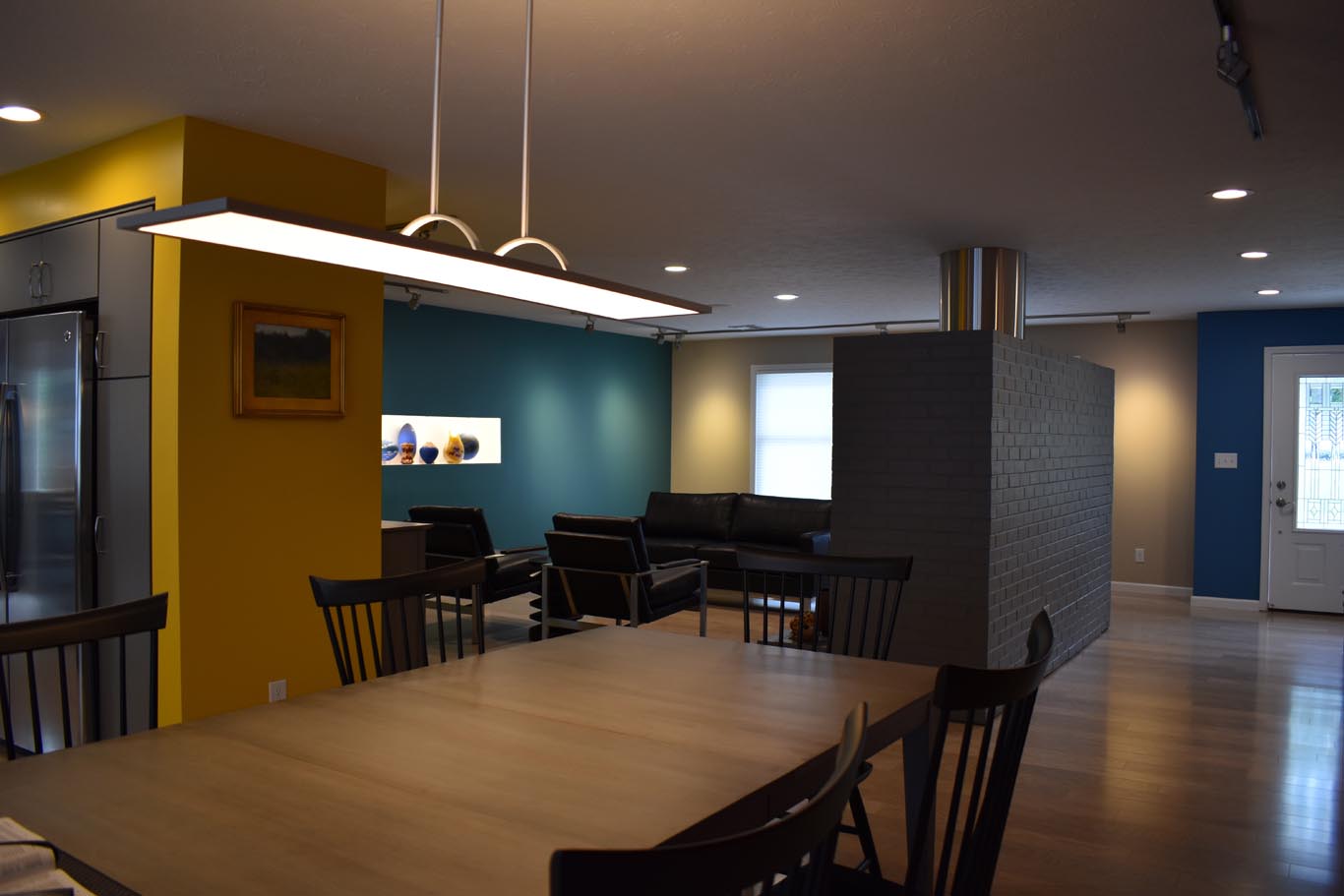 Modern open-concept living and dining area with wooden table, black chairs, sectional sofa, and accent walls in blue and yellow. Ceiling light fixture and wall art are visible.