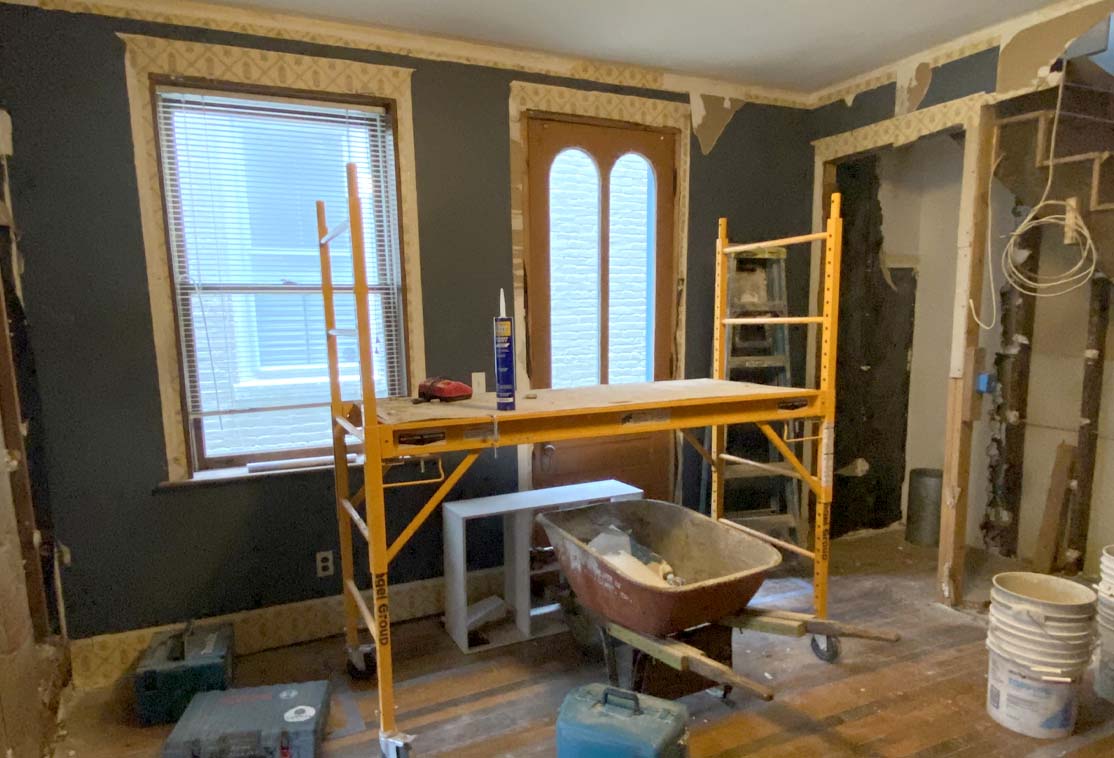 Room under renovation with scaffolding, a wheelbarrow, and construction tools. Walls are partially stripped, and materials are scattered on the floor.