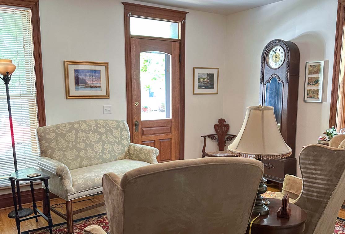 A cozy living room with upholstered chairs, a wooden grandfather clock, framed pictures on the walls, a floor lamp, and a wooden door with a window.