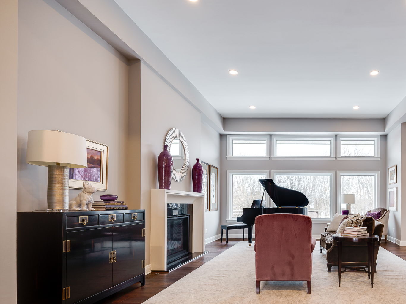 Spacious living room with a grand piano, armchairs, wooden floor, and bookshelves. Large windows illuminate the space, revealing elegant decor and vases on the shelves.