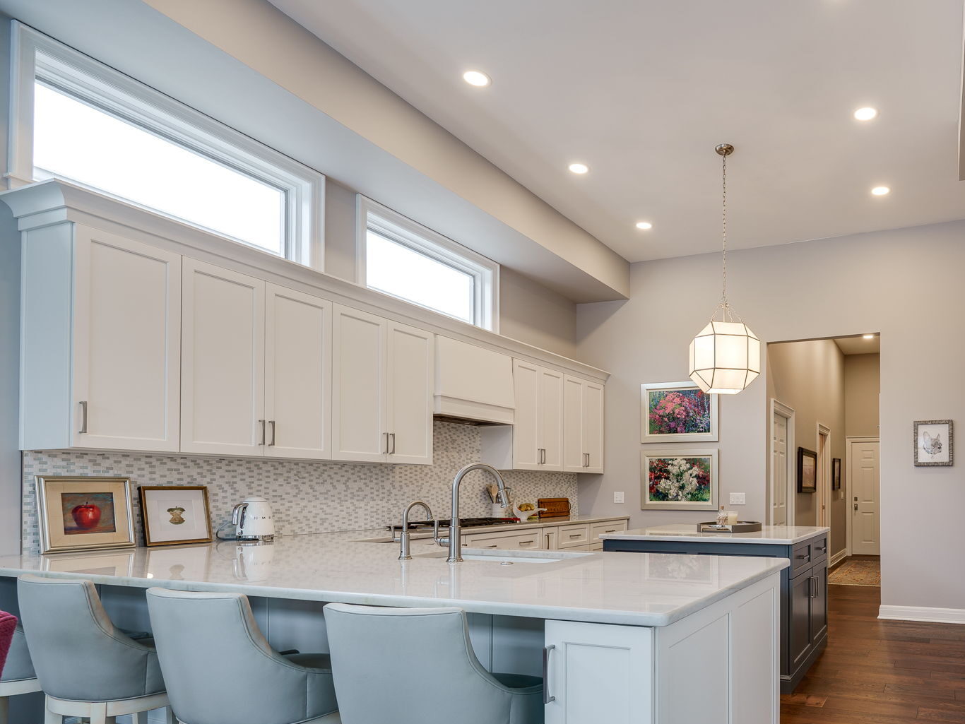 Modern kitchen with white cabinets, a central island with gray chairs, stainless steel appliances, and a wood floor. Pendant light hangs above the island. Artwork is displayed on the far wall.