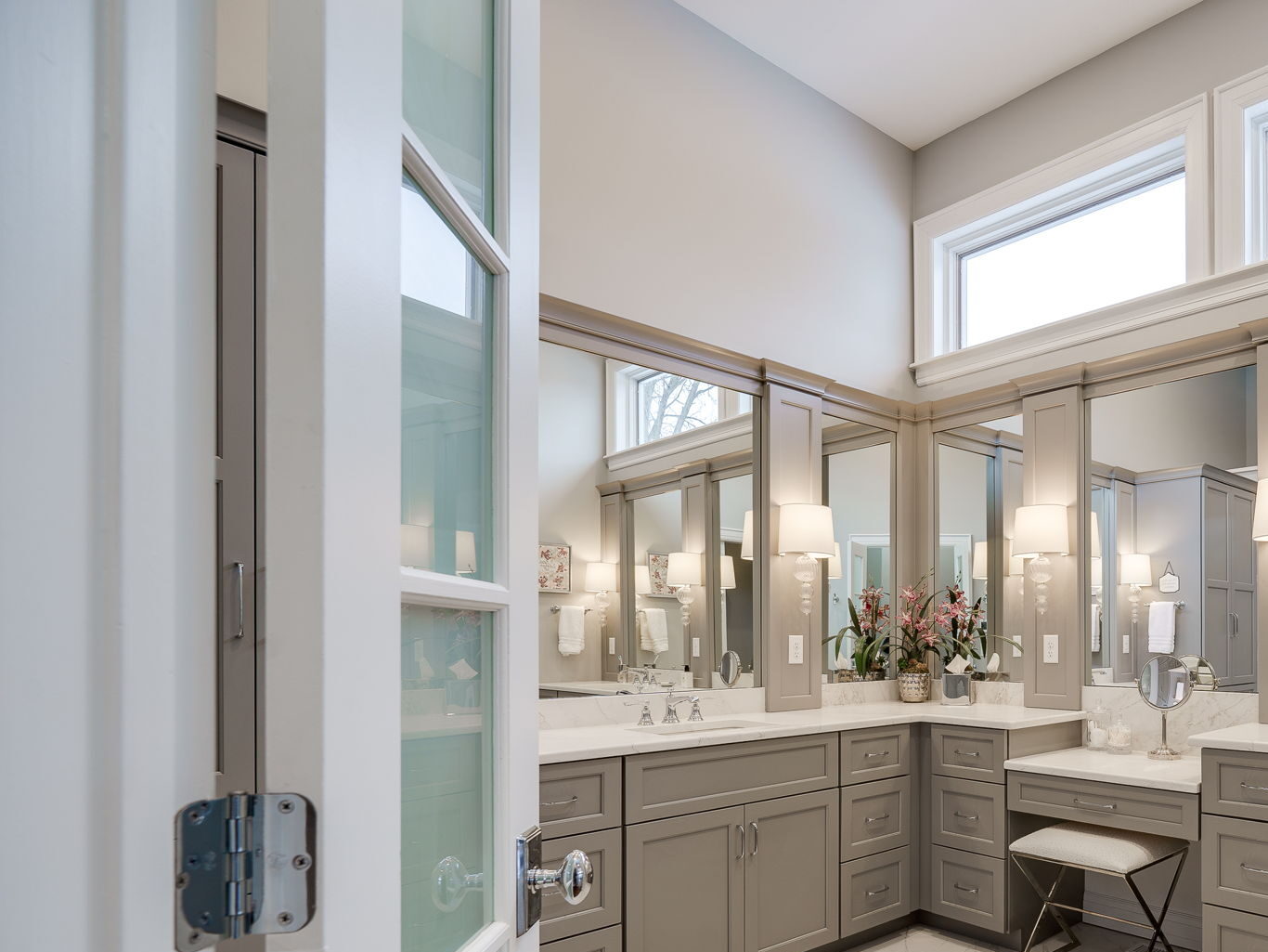 Spacious bathroom with double vanity, large mirrors, gray cabinets, and white marble floor. French doors open into the room, and a small stool is placed by the counters.