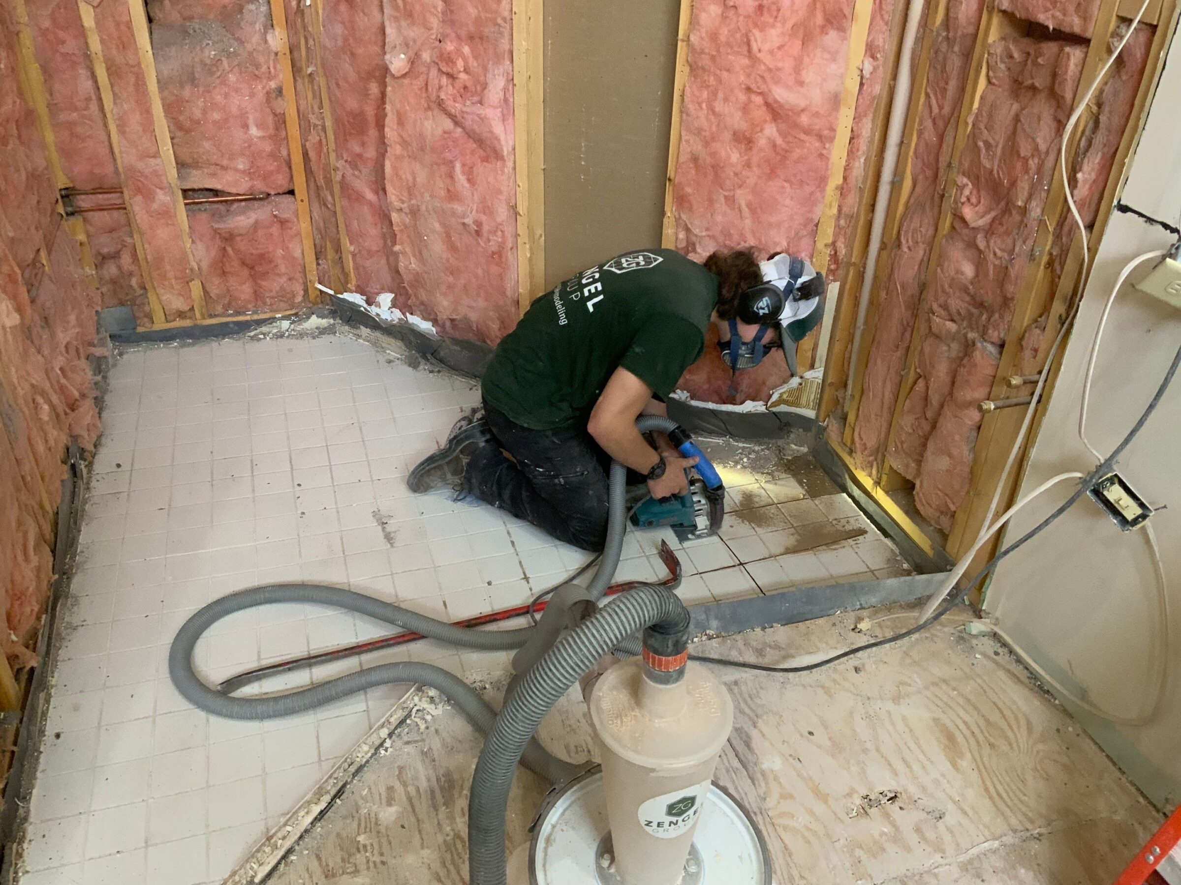 A worker kneels on a partially demolished bathroom floor, using a power tool with a hose and wearing safety gear; insulation and exposed wall studs are visible.