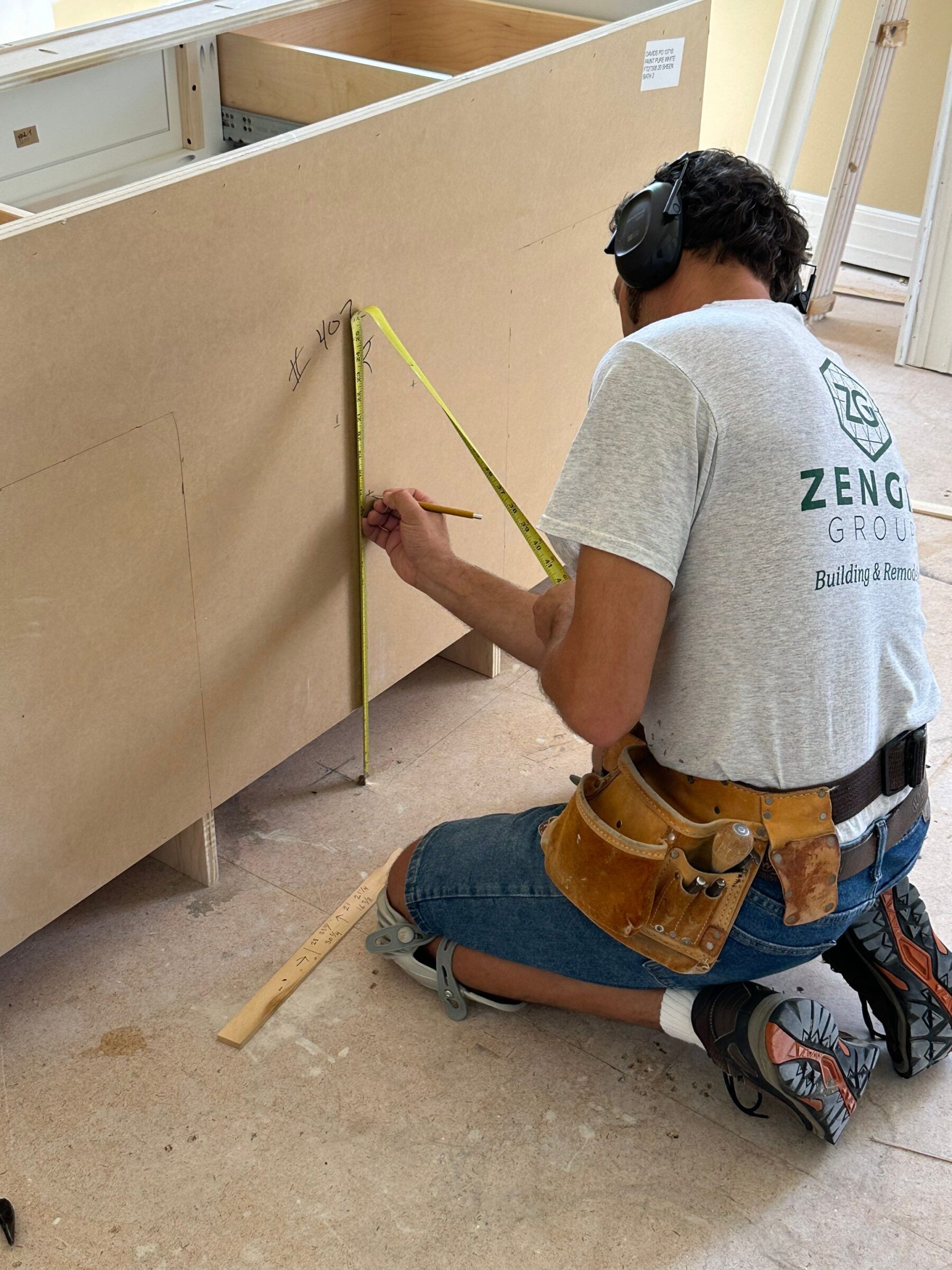 A worker wearing a tool belt and ear protection kneels on the floor, measuring and marking a cabinet panel with a tape measure and pencil.