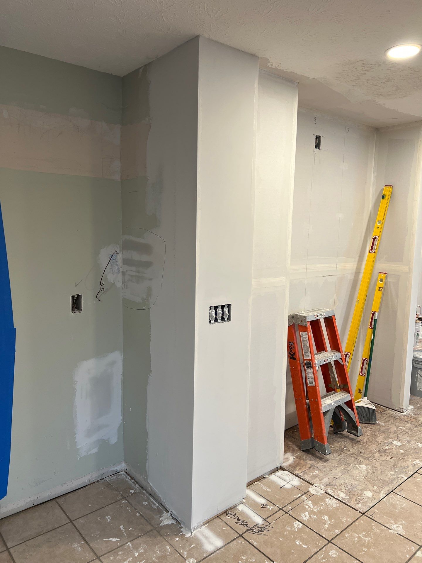 A partially renovated room with patched and primed drywall, a ladder, a level, and construction materials on a tiled floor.