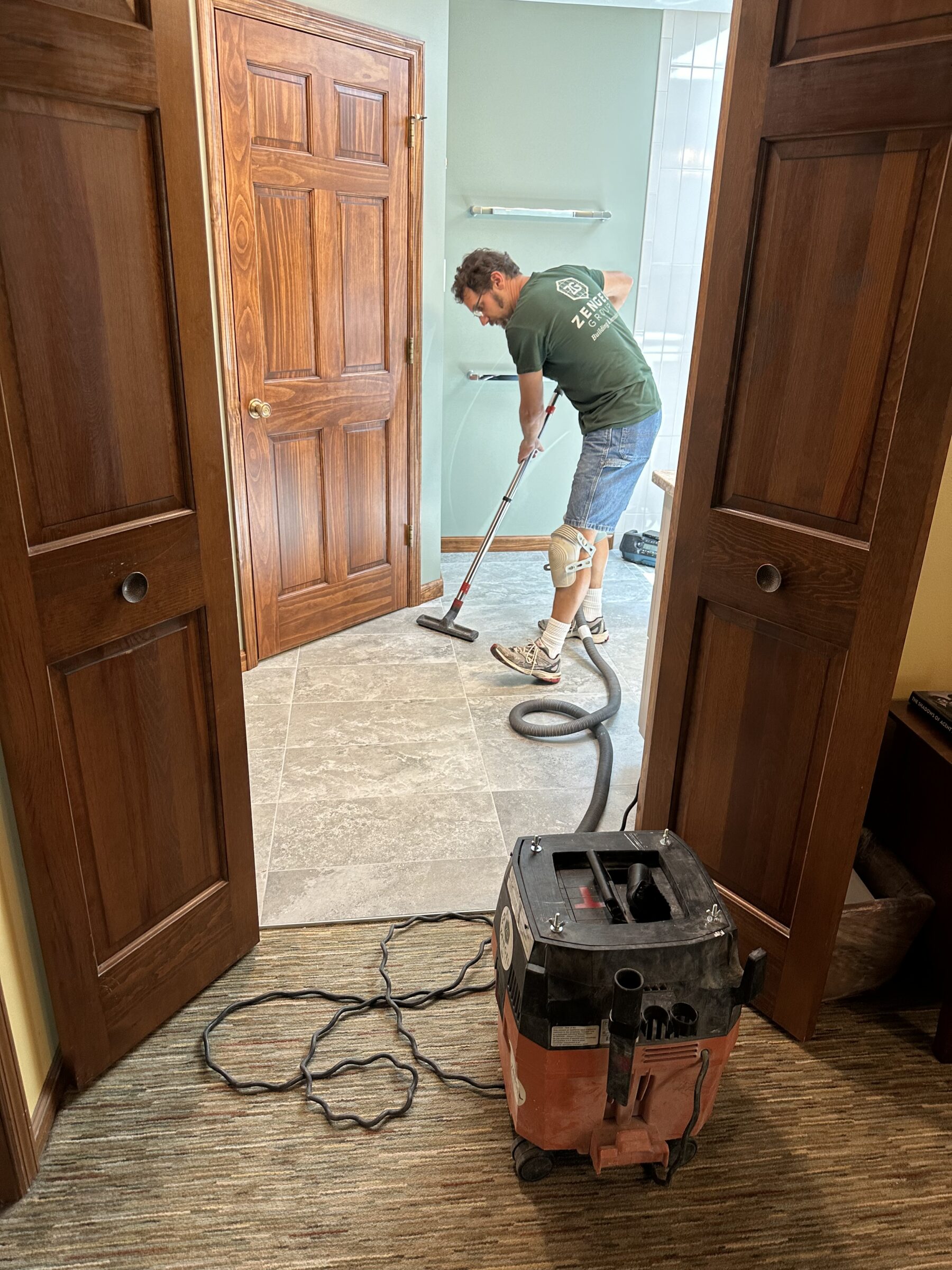 A man in work clothes uses a vacuum cleaner to clean a tiled floor in a room with wooden doors and light green walls.