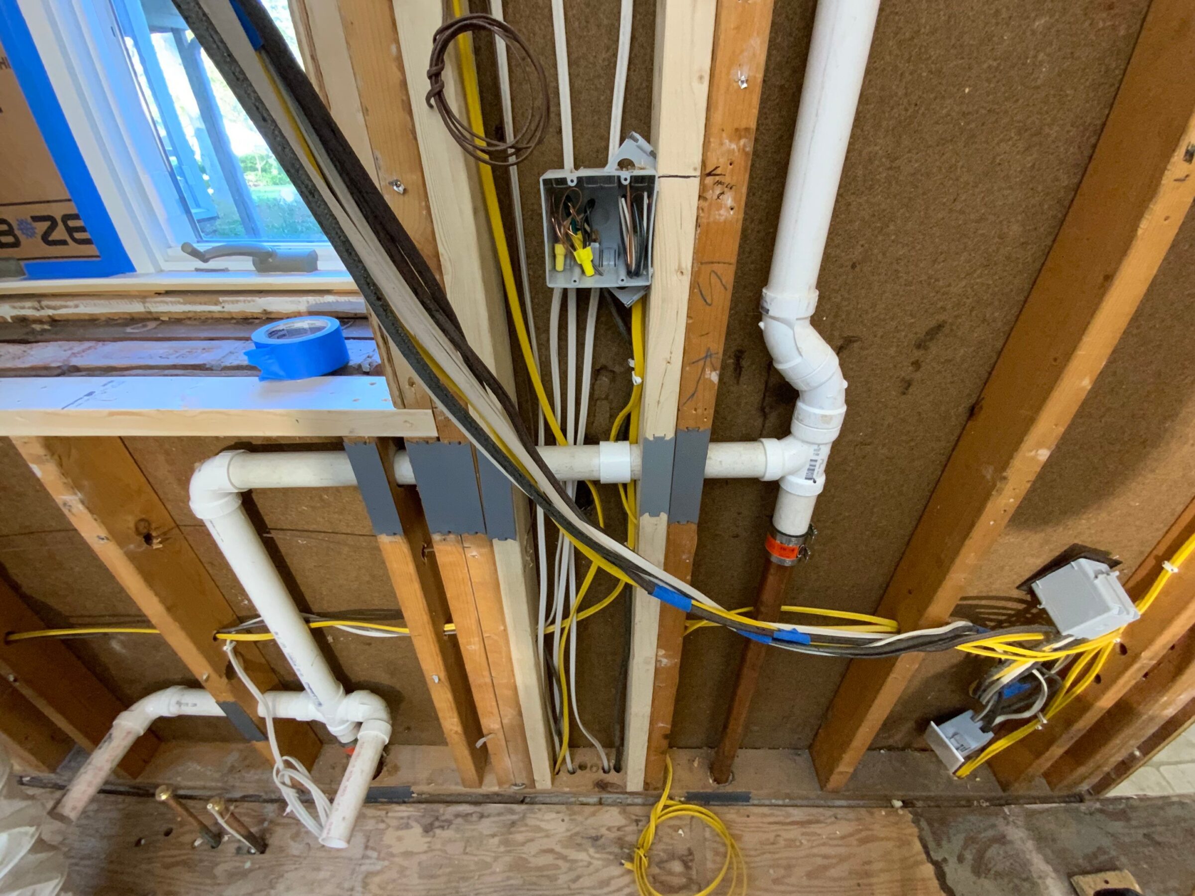 Exposed plumbing pipes and electrical wiring inside wall framing of a building under construction or renovation, with outlets, switch boxes, and window visible.