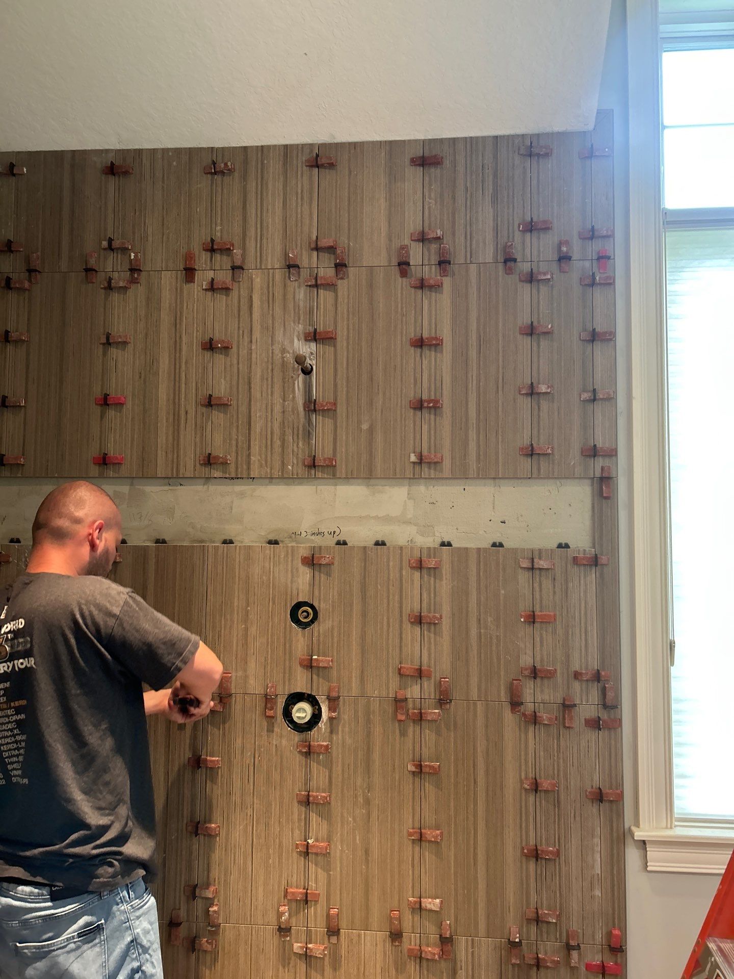 A person installs large rectangular tiles on a bathroom wall, using red tile spacers; plumbing fixtures are visible, and sunlight comes through a nearby window.