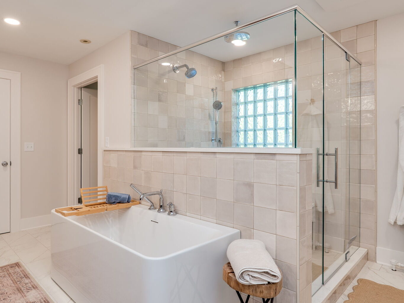 Modern bathroom with a freestanding tub, glass-enclosed shower, white walls, tiled floor, door, bathrobe on hook, and a stool with a folded towel beside the tub.