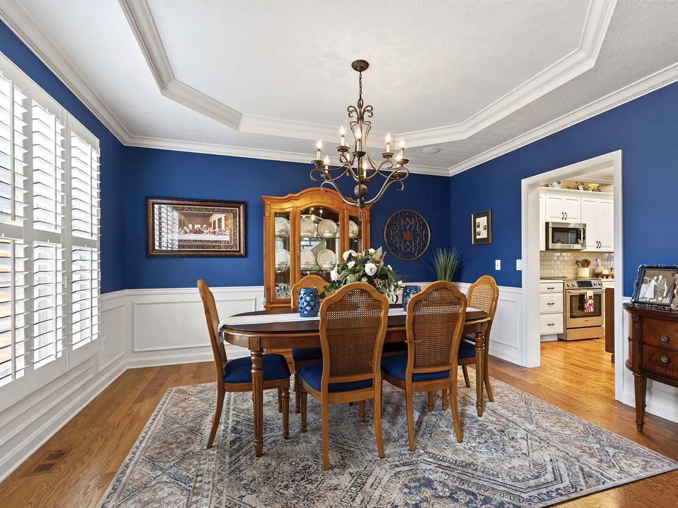 A formal dining room with blue walls, a wood table with six chairs, a chandelier, a china cabinet, and connecting view to a white kitchen.