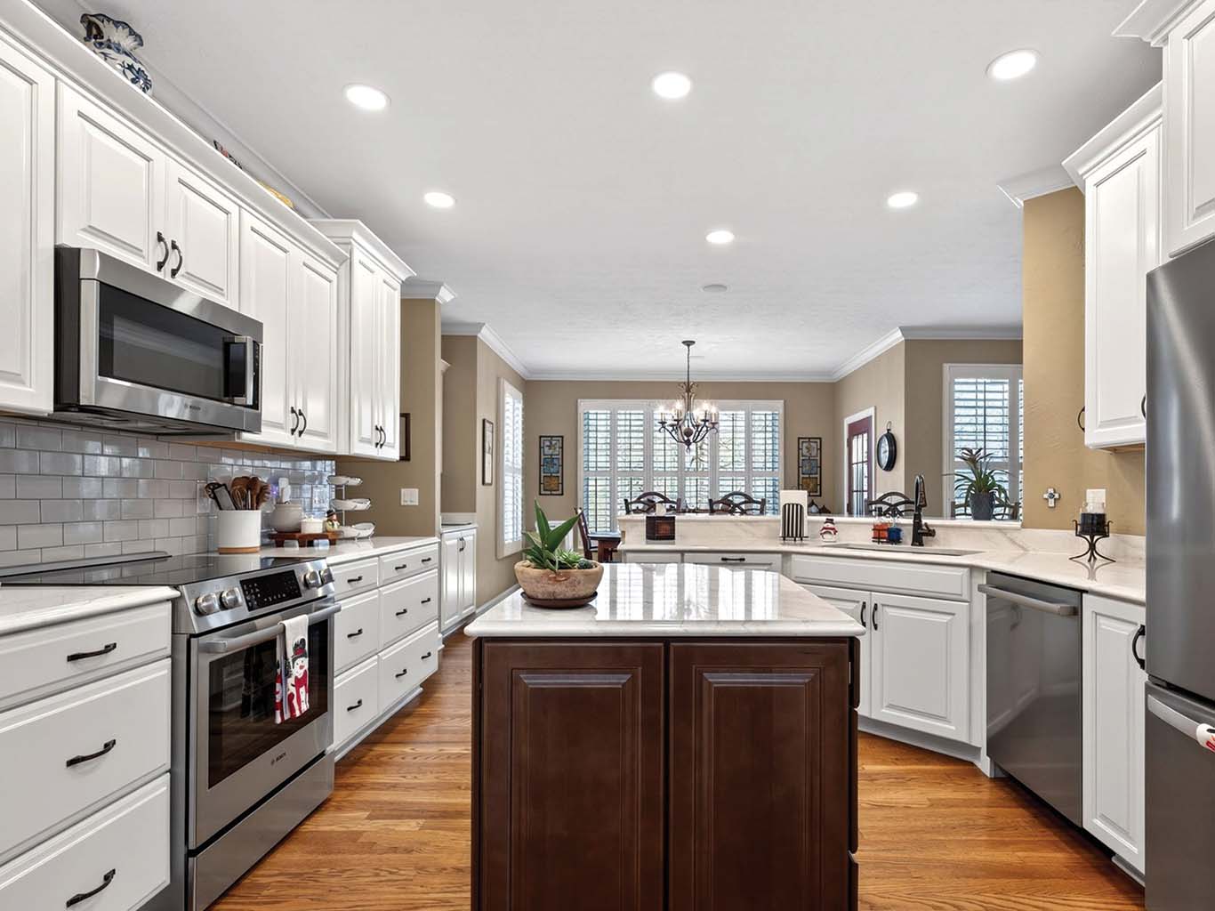 Modern kitchen with white cabinets, stainless steel appliances, dark wood island, hardwood floors, and recessed lighting; dining area visible in the background.