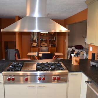 Modern kitchen with a stainless steel range hood, gas stove, light cabinetry, dark countertops, and a mosaic tile backsplash. Dining area visible in the background.