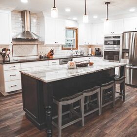 A modern kitchen with a center island, stools, stainless steel appliances, white cabinets, and wooden floors. Three pendant lights hang above the island.