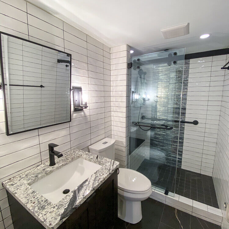 A modern bathroom features a granite countertop sink, toilet, and a glass-enclosed shower. Walls are tiled in white with black grout.