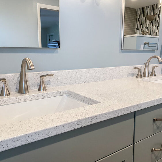 Modern bathroom with dual sinks, silver faucets, white speckled countertop, and light blue cabinets. A towel hangs on the wall to the right.